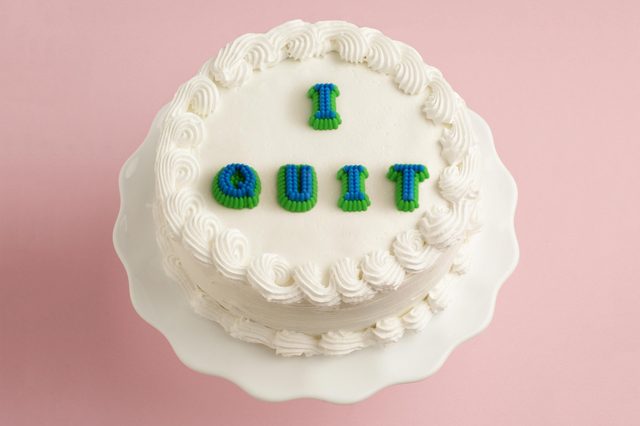 Cake displays "I QUIT" in blue and green icing, resting on a white stand against a pink background.