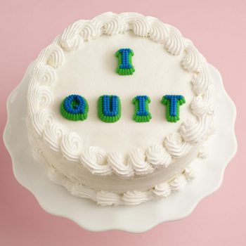 Cake displays "I QUIT" in blue and green icing, resting on a white stand against a pink background.