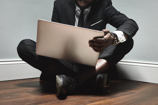 Man in suit sits cross-legged, holding laptop, on wooden floor against a light-colored wall.