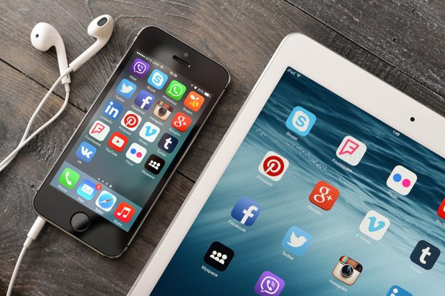 Smartphone displaying social media apps; headphones beside it; tablet nearby with similar icons; all on a wooden surface.