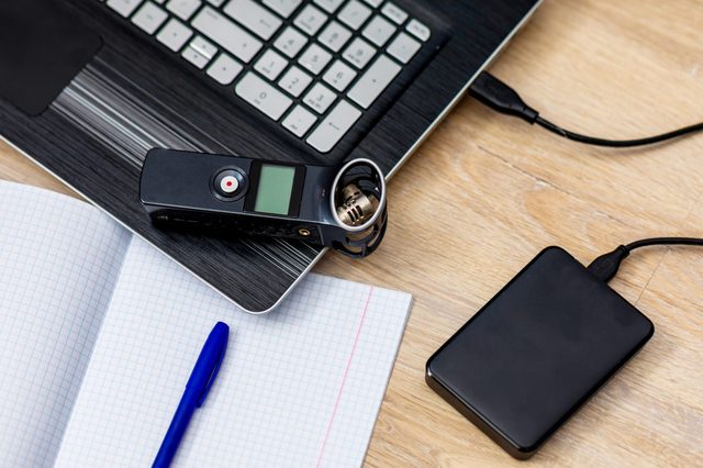 Voice recorder rests on a laptop beside a notebook and pen, with an external hard drive nearby on a wooden desk.