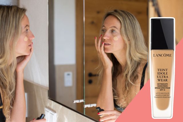 Woman applies foundation on her face, looking into a mirror in a bathroom. A Lancome Teint Idole Ultra Wear bottle appears on the side.