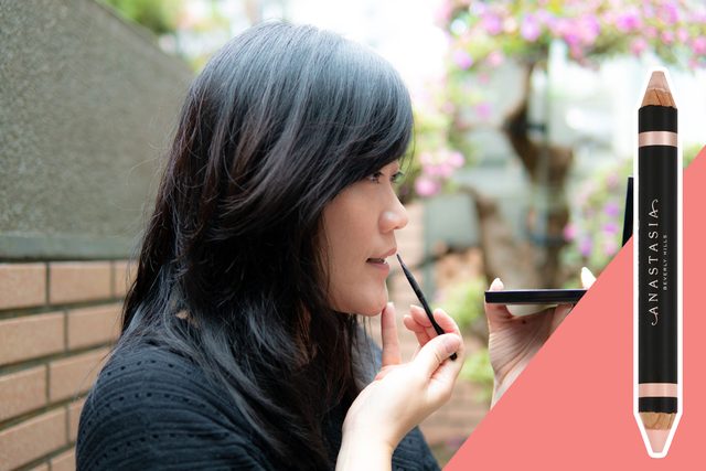 Woman applies makeup using a mirror, surrounded by an outdoor setting with greenery. Right side features an Anastasia Beverly Hills makeup pencil.