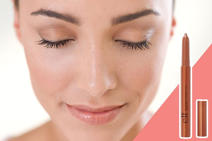 Woman closes eyes, showcasing natural makeup; beside her, an e.l.f. eye shadow stick is displayed on a pink background.