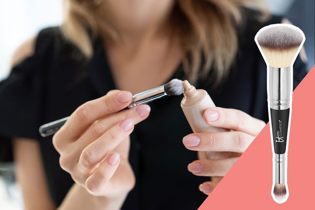 A person applies makeup using a brush, holding a small bottle of foundation, with a close-up view on a pink background.
