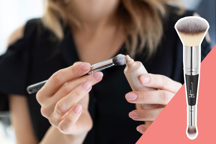 A person applies makeup using a brush, holding a small bottle of foundation, with a close-up view on a pink background.