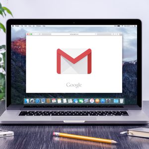 Laptop displaying Gmail logo sits on a desk with a smartphone, pencil, and notebook; plant and water cooler in the background.