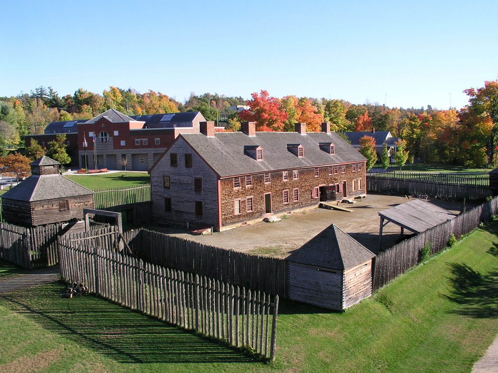 Wooden fort sits enclosed by fence, surrounded by autumn trees and modern buildings in background under clear blue sky.