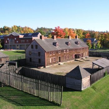 Wooden fort sits enclosed by fence, surrounded by autumn trees and modern buildings in background under clear blue sky.