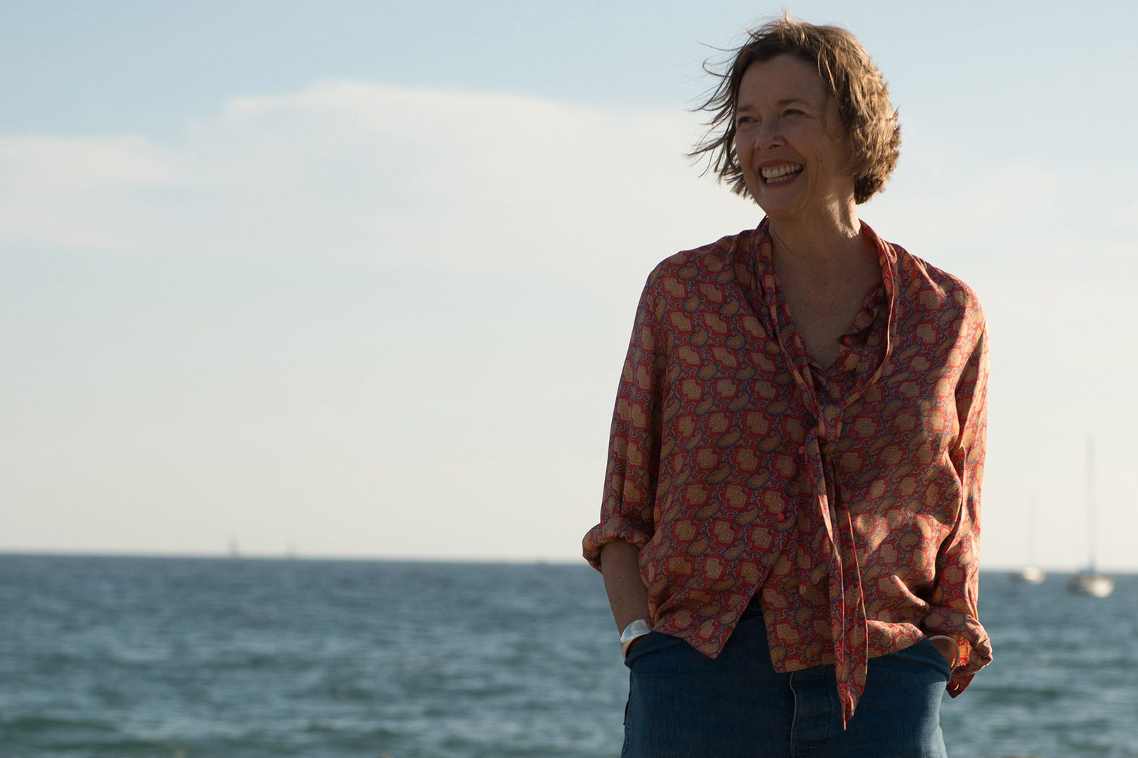 A person smiles broadly, standing by the ocean with sailboats in the background under a clear sky.
