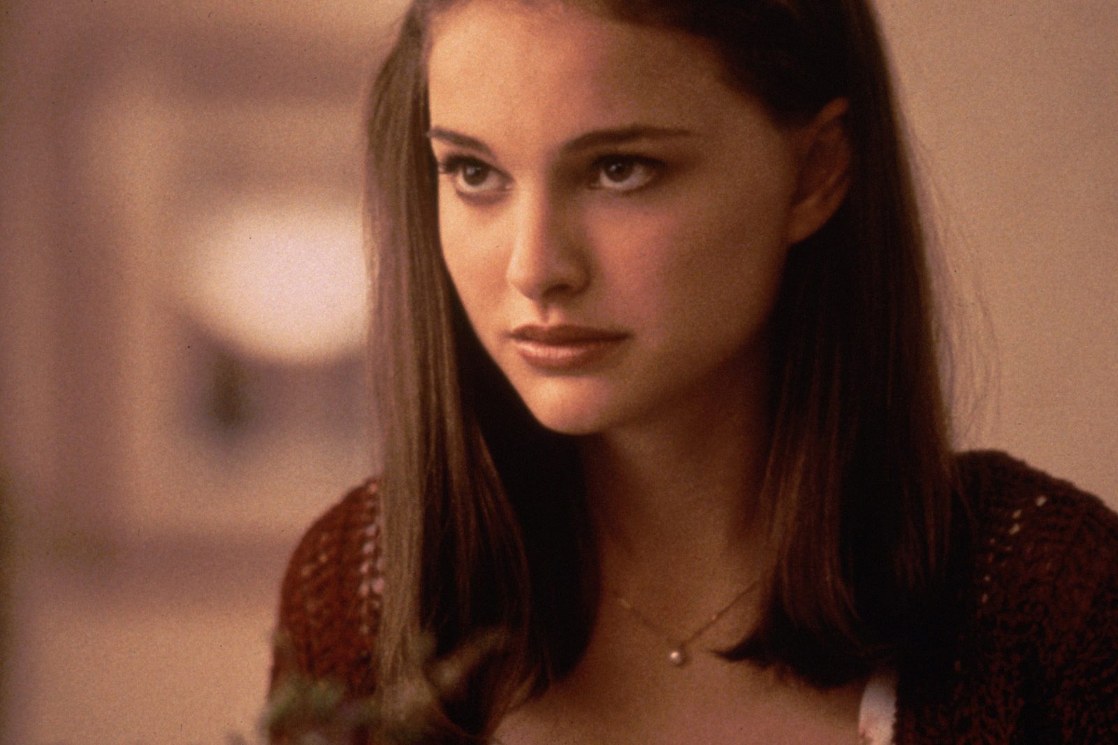 A woman stares intently, wearing a brown sweater and necklace, set against a blurred indoor background.