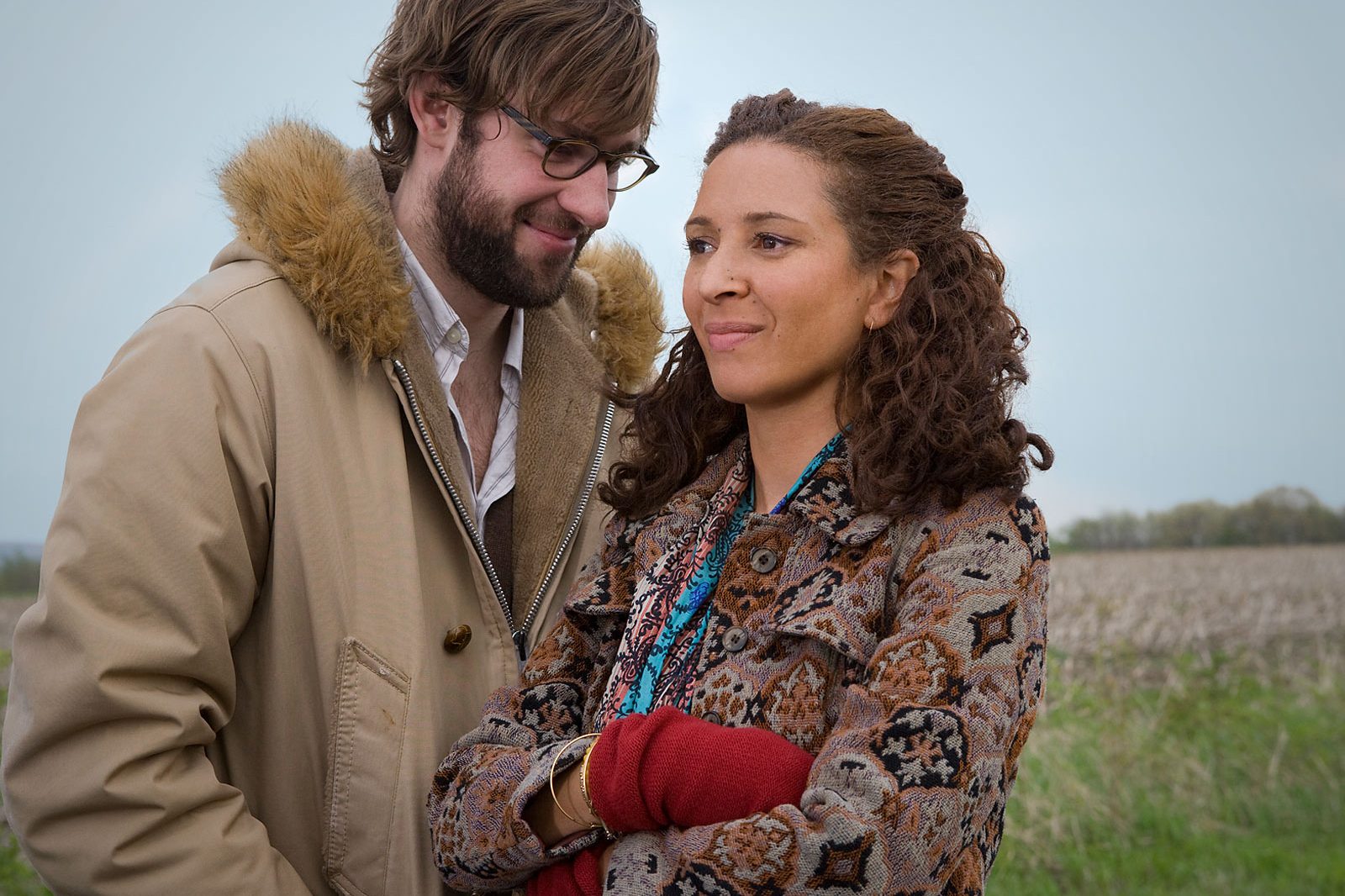 Two people stand together, one smiling at the other. They're in a field, wearing warm, patterned clothing. The scene suggests a relaxed conversation outdoors.