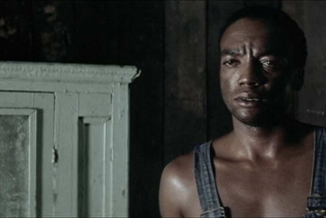 A man stands, wearing overalls, looking thoughtful in a dimly lit room with wooden walls and a white wooden cabinet.