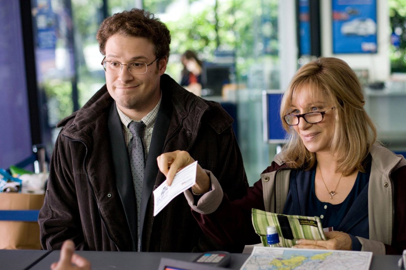 Two people stand at a counter, handing over a document in an office setting with maps and brochures visible.