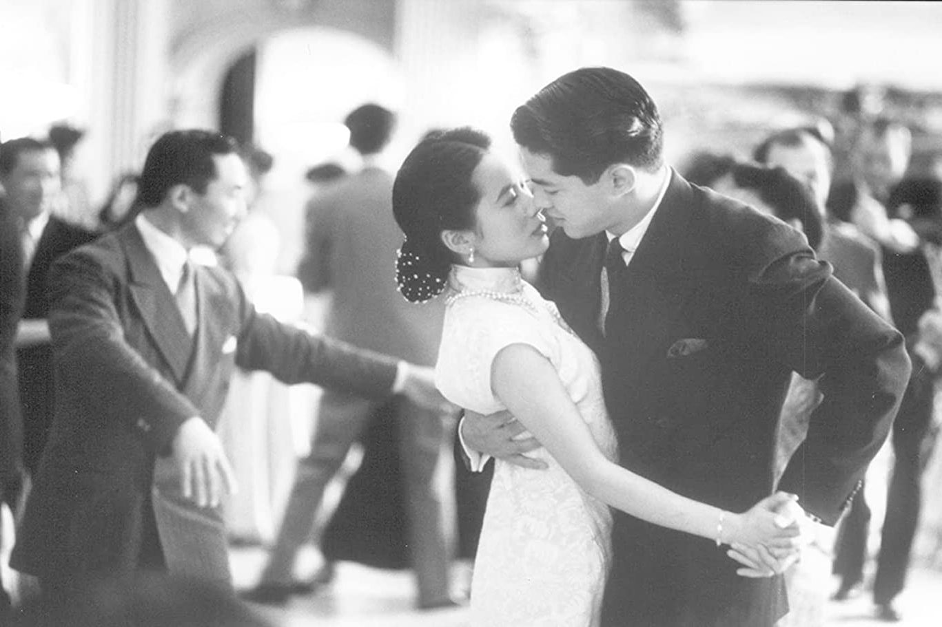 A dancing couple embraces intimately in a crowded ballroom, surrounded by other elegantly dressed people.