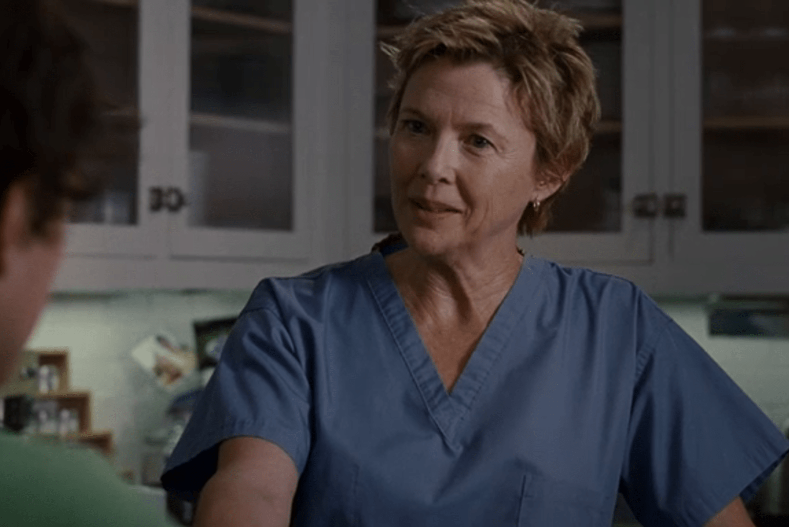 Person wearing blue scrubs is speaking, standing in a kitchen with white cabinets.