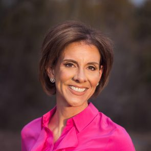 A person smiles while wearing a bright pink blouse; the background is blurred and natural, possibly outdoors.