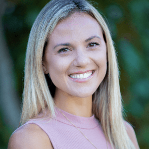 A person smiles outdoors, framed by green foliage. They wear a pink top and a necklace with the name "Allie."