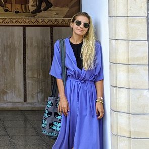 Woman leans against a stone wall, wearing a purple dress and sunglasses, holding a blue-patterned bag. Mural art behind her.