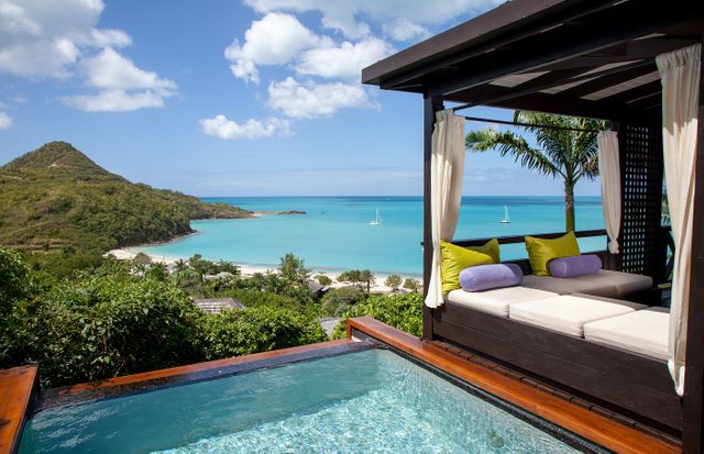 Lounge bed overlooks infinity pool and turquoise bay with sailboats, nestled in lush, tropical setting under a clear blue sky.