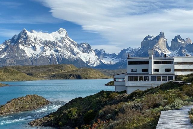 A building overlooks a turquoise lake with a backdrop of snow-capped mountains under a partly cloudy sky.