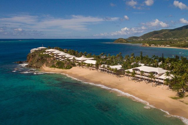 Beachfront resort overlooks turquoise ocean, surrounded by palm trees. White-roofed buildings line the sandy shore under a clear blue sky.