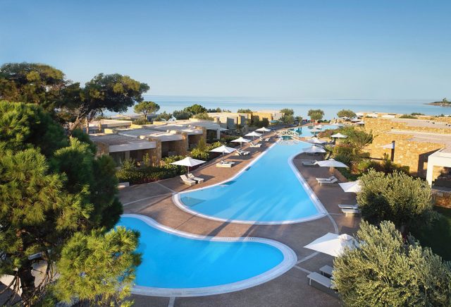A winding pool reflects sunlight, surrounded by lounge chairs with umbrellas, situated in a resort complex near a calm ocean horizon.