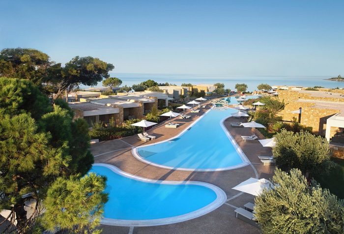 A winding pool reflects sunlight, surrounded by lounge chairs with umbrellas, situated in a resort complex near a calm ocean horizon.