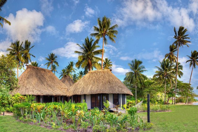 Thatched-roof huts stand amid vibrant tropical greenery, surrounded by tall palm trees and a bright blue sky.