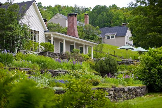 A white house sits peacefully, surrounded by vibrant gardens with purple flowers and greenery on a lush, terraced hillside under a clear sky.