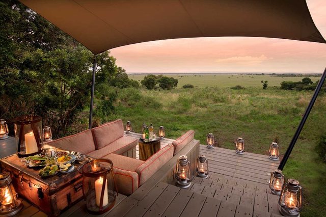 Plush sofas overlook a scenic savanna, with a spread of food on a wooden table, surrounded by lanterns under a canopy.