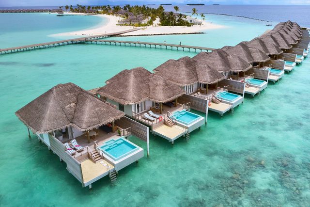 Overwater bungalows stand above turquoise water, connected by wooden walkways, with a distant white sand beach lined with palm trees.