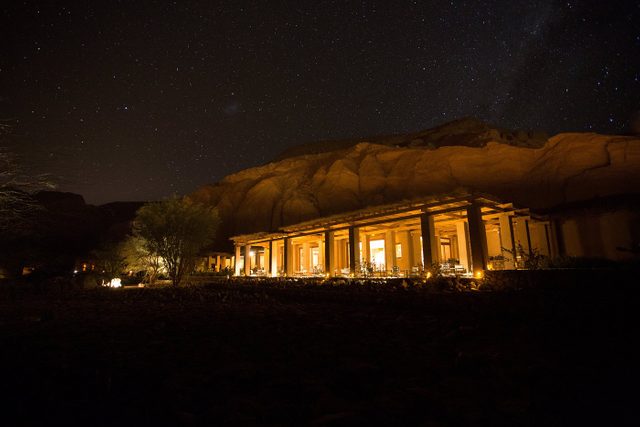 Illuminated building rests under starry night sky, set against rugged, rocky hills. Sparse trees and shrubs surround, casting faint shadows in the foreground.