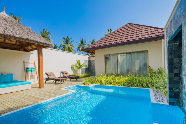 Pool reflecting sunlight, chairs and cabana nearby, surrounded by lush greenery against a clear blue sky.