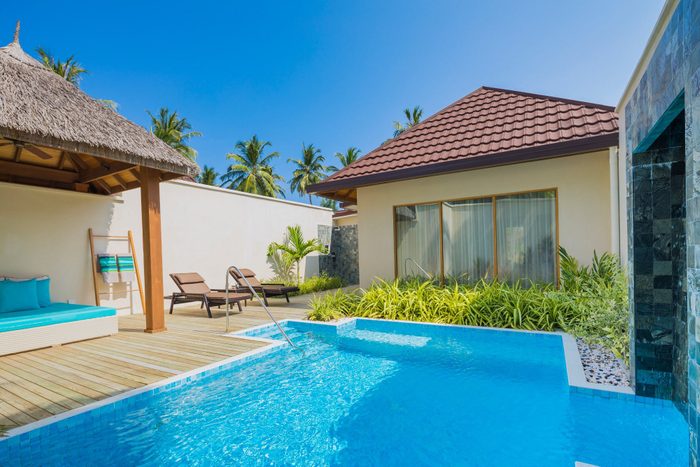 Pool reflecting sunlight, chairs and cabana nearby, surrounded by lush greenery against a clear blue sky.