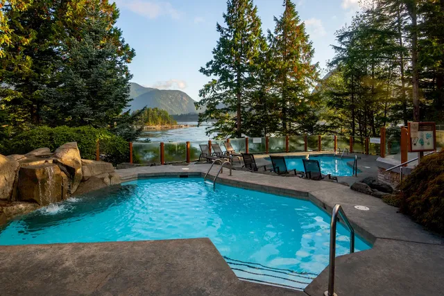 A swimming pool sits quietly surrounded by trees and mountains, with lounge chairs arranged nearby under a clear sky.
