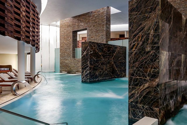 Indoor pool flowing gracefully in a modern spa featuring stone walls and lounge chairs, creating a serene, luxurious ambiance.
