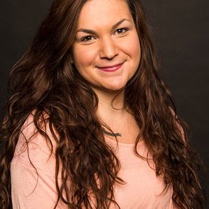 A person sits smiling against a plain, dark background, wearing a pink top.