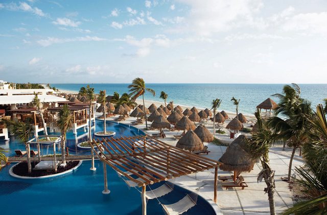Poolside loungers face a sunlit ocean, surrounded by palm trees and straw huts, creating a tropical resort atmosphere.