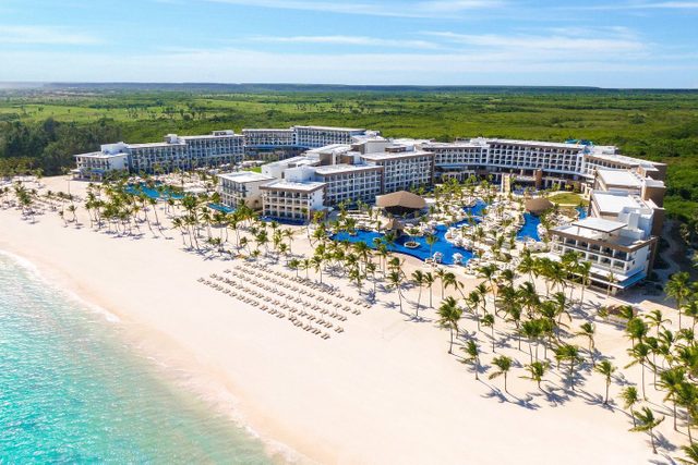 Resort stretches along sandy beach, bordered by turquoise water and lush greenery, with rows of palm trees and lounge chairs.