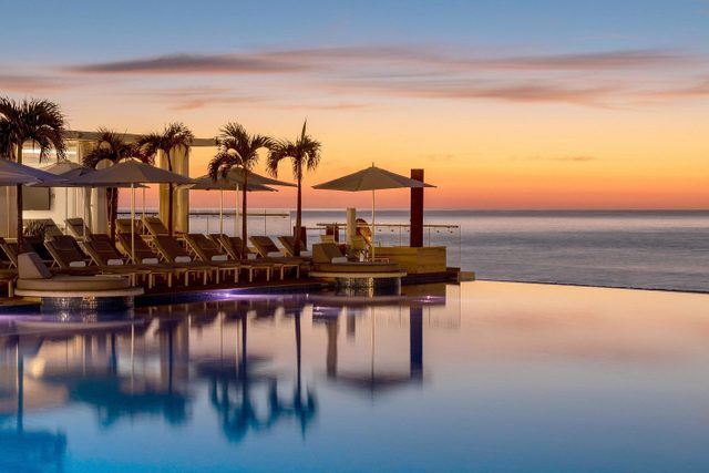 Lounge chairs sit under umbrellas beside a pool, reflecting a vibrant sunset, with the ocean as a backdrop.