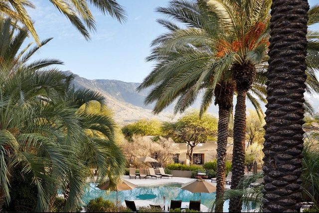 Palm trees surround a tranquil pool with lounge chairs and umbrellas, set against a backdrop of mountains under a clear blue sky.