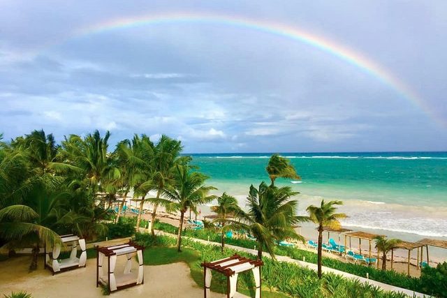 Rainbow arches over turquoise ocean, bordered by lush palm trees and canopied beach beds under a cloudy sky.