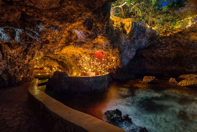 Table with chairs, adorned with candles in a rocky cave, set beside a gently lit, serene pool.
