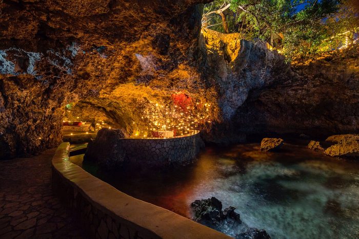 Table with chairs, adorned with candles in a rocky cave, set beside a gently lit, serene pool.
