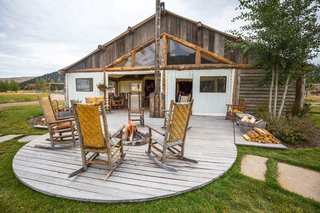 Wooden rocking chairs encircle a small fire pit on a wooden deck, outside a rustic lodge, surrounded by greenery and mountains.