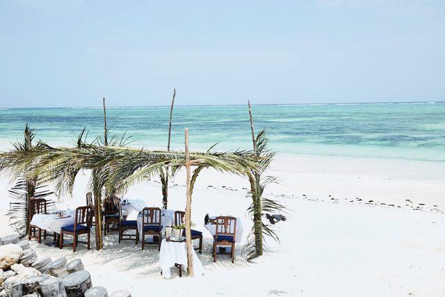 Dining table set under palm frond canopy, overlooking a clear, turquoise beach with white sand and serene ocean views.