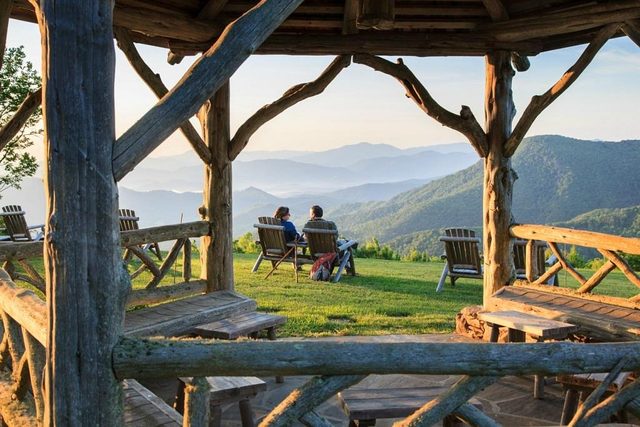 Chairs face mountains; people sit on grass under a wooden gazebo, overlooking misty hills at sunset.