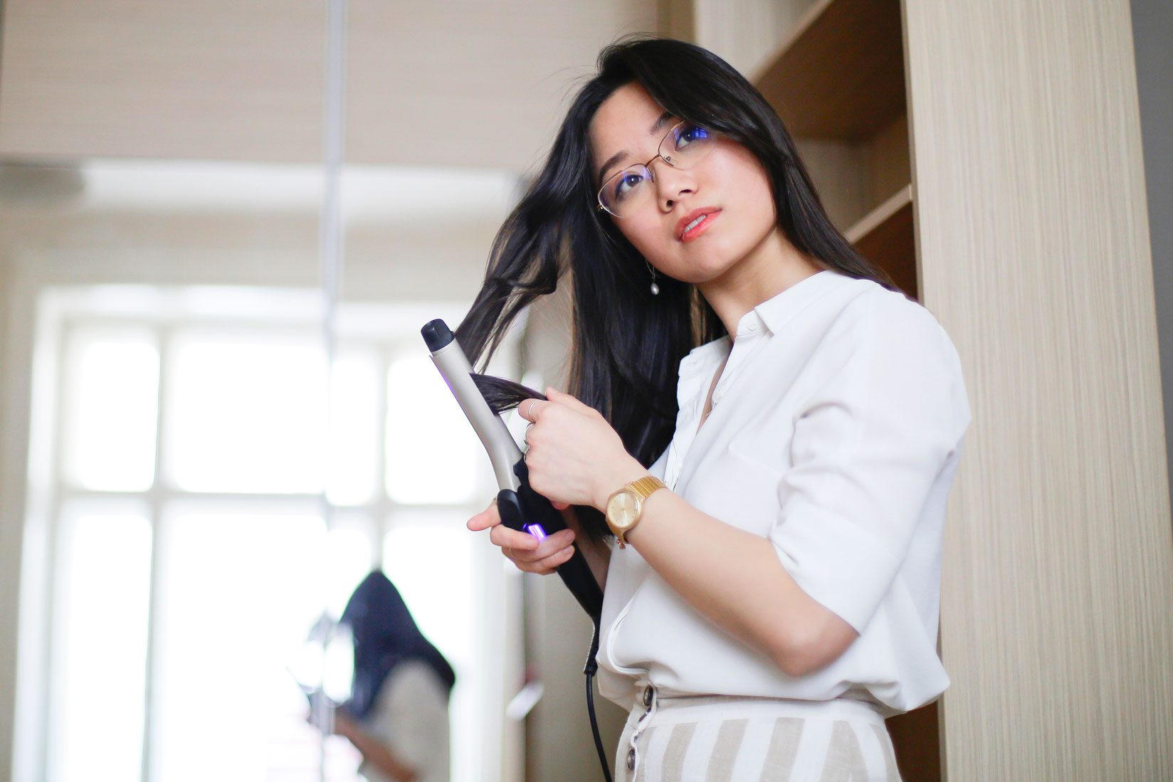 A person uses a curling iron on their hair in a bright room with a window and mirror in the background.