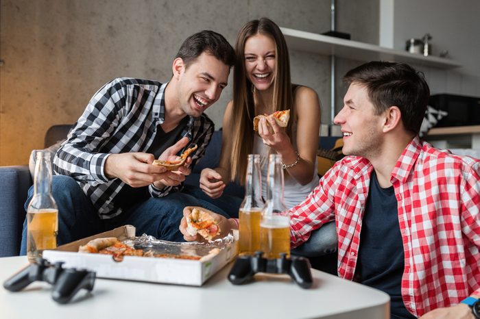 happy young people eating pizza, drinking beer, having fun, friends party at home, hipster company together, two men one woman, smiling, positive, relaxed, hang out, laughing, funny emotions
